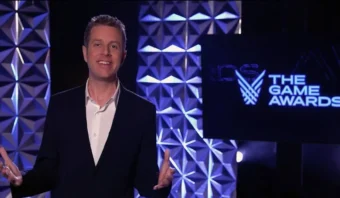 Geoff Keighley con traje se encuentra en el escenario frente a un cartel de "The Game Awards", hablando y gesticulando con las manos, rodeada de una iluminación geométrica.