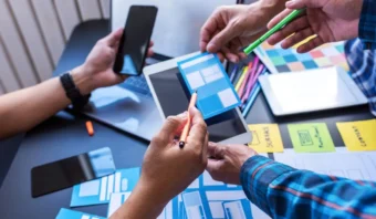 En una mesa, las personas que trabajan juntas con smartphones, tablets y diseños en papel sostienen bolígrafos y discuten planes de diseño. Su conversación refleja los principios de Ley de Jakob, que enfatizan una experiencia de usuario intuitiva alineada con patrones familiares.