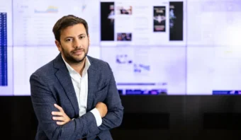 Una persona con una chaqueta azul y una camisa blanca está de pie con los brazos cruzados frente a una pantalla que muestra gráficos y artículos.