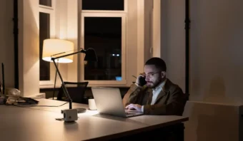 Un hombre trabaja con una computadora portátil en un escritorio en una habitación poco iluminada, con un teléfono en la oreja y una lámpara iluminando el espacio.