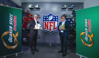 Dos hombres están de pie frente a un fondo con el logotipo de la NFL, cada uno de ellos sosteniendo objetos. En la pared se exhiben cascos de la NFL, flanqueados por carteles de Quaker State que añaden un toque de estilo patrocinador a la escena.