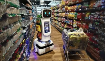 Un Shopperbot recorre el pasillo del supermercado, rodeado de estantes con bocadillos y bebidas, junto a un carrito de compras repleto de artÃ­culos.