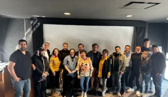 Un grupo de personas posa para una foto en una sala frente a una gran pantalla que muestra el logotipo de AMFI 2025-2026. La sala, con su interior oscuro, está sutilmente iluminada por la luz que entra a través de una ventana a la izquierda.