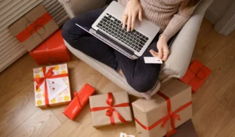 Persona sentada en una silla usando una computadora portátil con una tarjeta de crédito en la mano, rodeada de regalos envueltos en el suelo.