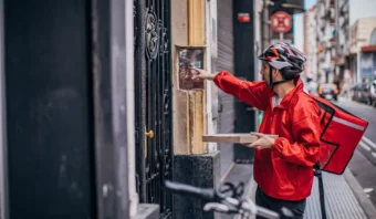 Un repartidor con chaqueta roja y casco presiona un timbre mientras sostiene una caja de pizza y hay una bicicleta estacionada cerca en una calle de la ciudad.