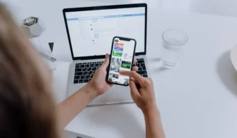 Persona navegando en un teléfono inteligente mientras está sentada en un escritorio con una computadora portátil abierta y un vaso de agua.