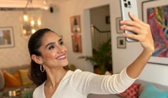 Una mujer con una camisa blanca de manga larga se toma una selfie en una sala de estar cálidamente iluminada con una decoración colorida.