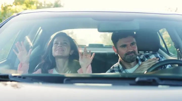 Una mujer y un hombre están sentados en un automóvil, atrapados en medio del tráfico, mirando hacia adelante con las manos levantadas hacia el parabrisas.