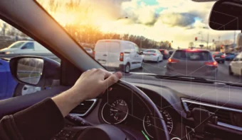 Vista desde el interior de un automóvil que muestra la mano de un conductor en el volante, rodeado de tráfico, con un cielo de atardecer en el fondo.
