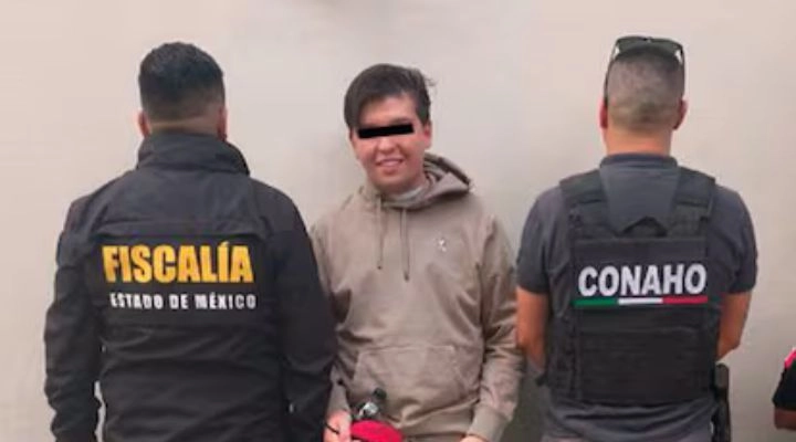 Una persona se encuentra de pie entre dos oficiales, uno con una chamarra de “Fiscalía Estado de México” y el otro con un chaleco de “Conaho”. La persona está mirando hacia la cámara, mientras los oficiales miran hacia otro lado.