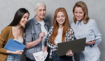 Cuatro mujeres de distintas edades están juntas, sonriendo y mirando una computadora portátil. Una sostiene un portapapeles, otra sostiene papeles con gráficos. Están sobre un fondo gris liso.