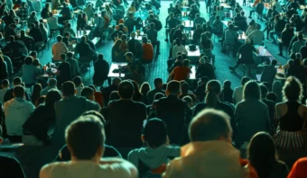 Una gran multitud de personas sentadas al aire libre por la noche, mirando hacia una dirección común. Las mesas están dispersas entre los asistentes. La iluminación sugiere un evento o una reunión organizada.