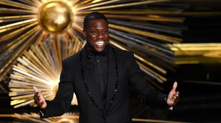 Kevin Hart sonríe y gesticula mientras está de pie frente a un fondo dorado con estrellas en Los Oscars