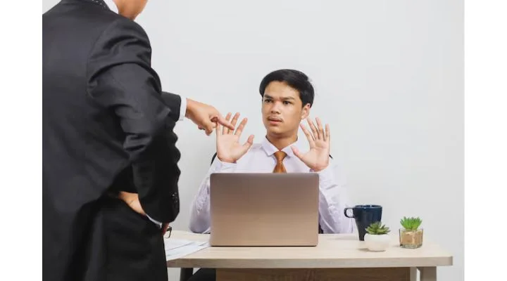 Una persona sentada en un escritorio con una computadora portátil levanta las manos en defensa mientras otra persona vestida de traje señala y habla.