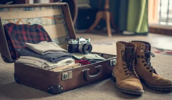 Una maleta abierta revela ropa doblada y una cámara en su interior, combinada con un toque elegante de estilo retro-vintage: unas botas marrones apoyadas en el suelo completan este conjunto de viaje atemporal.