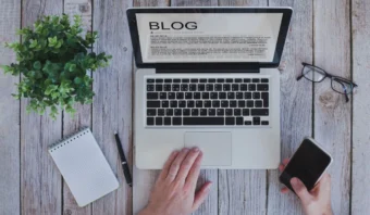 Una persona escribe en un portátil que muestra un blog. Sobre una mesa de madera hay un teléfono inteligente, unas gafas, un cuaderno y una maceta con una planta.