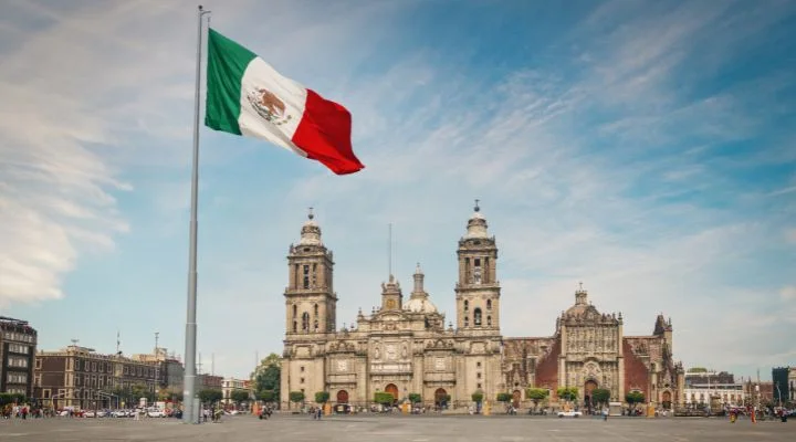 Una gran bandera mexicana ondea en la Plaza del Zócalo, Ciudad de México, con la Catedral Metropolitana al fondo bajo un cielo parcialmente nublado, capturando la esencia que Emilia Pérez a menudo buscaba retratar en sus vibrantes obras de arte.