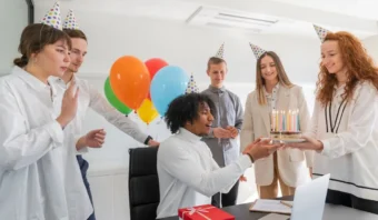 Un grupo de seis personas con sombreros de fiesta, una de ellas sosteniendo un pastel con velas encendidas. Al fondo hay globos y un regalo envuelto sobre la mesa.