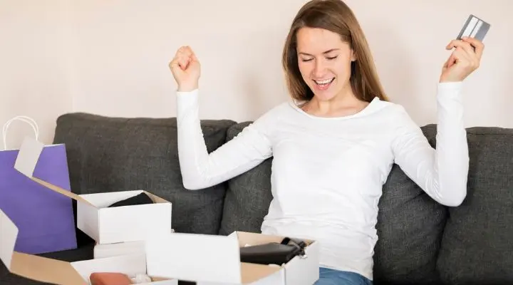 Una mujer sentada en un sofá, sosteniendo una tarjeta de crédito y levantando el puño con entusiasmo en medio de la emoción del unboxing, rodeada de cajas de compras abiertas.