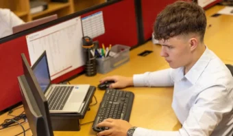 Persona con camisa blanca trabajando en un escritorio con una computadora portátil, un teclado y un monitor. Los útiles de oficina están organizados cerca.