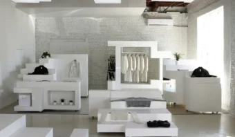 Interior de boutique minimalista y moderno con exhibidores geométricos blancos, percheros y pequeñas plantas en macetas. La luz natural entra por los amplios ventanales, iluminando el espacio.