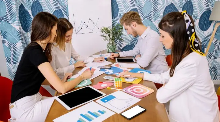 Cuatro personas del departamento de marketing se sientan en una mesa con tablets e iconos de redes sociales, discutiendo estrategias y anotando ideas. Un grÃ¡fico en la pared, detrÃ¡s de ellas, complementa su anÃ¡lisis.