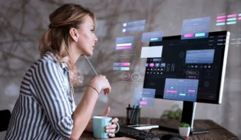 Mujer trabajando en una computadora con pantallas de interfaz holográficas, sosteniendo un bolígrafo y una taza.