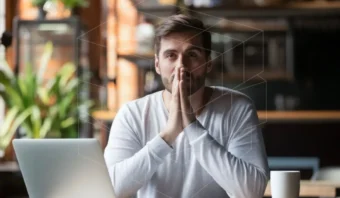 Un hombre se sienta en una mesa con una computadora portátil y una taza de café, apoyando sus manos en su rostro y mirando hacia adelante en un ambiente interior luminoso.
