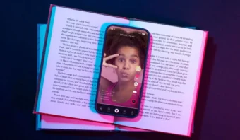 Teléfono inteligente que muestra a una persona haciendo el signo de la paz en una aplicación de video, apoyada sobre un libro abierto con iluminación de colores.