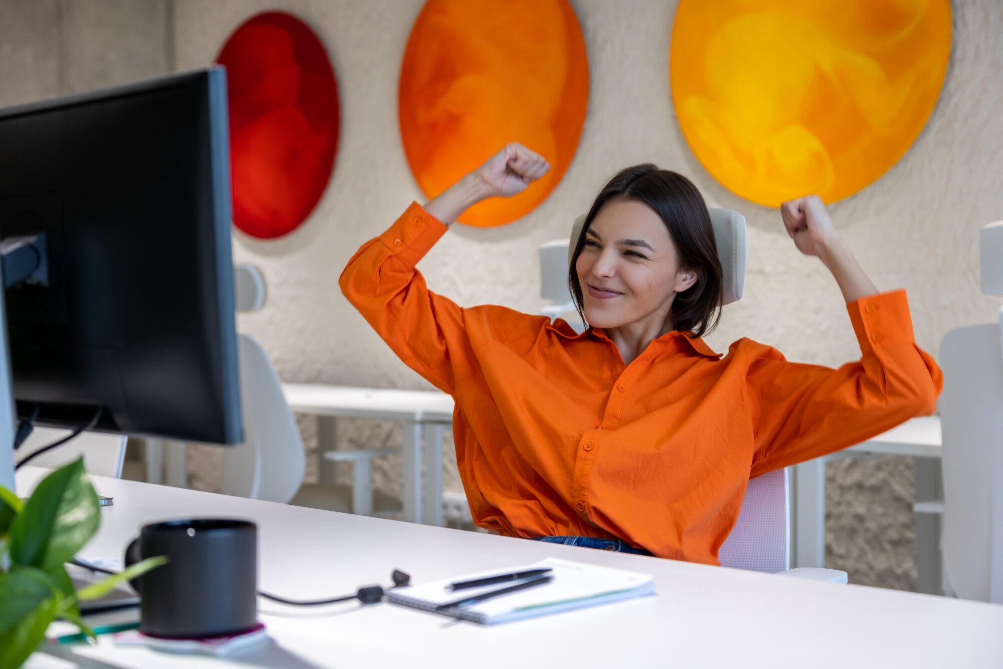 Una mujer con una camisa naranja está sentada en un escritorio, estirándose y sonriendo frente a un monitor de computadora, con arte circular colorido en la pared detrás de ella.