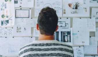 Una persona con cabello castaño corto y un suéter a rayas mira una pared cubierta de papeles, gráficos y diagramas dispuestos para realizar una lluvia de ideas o planificar.