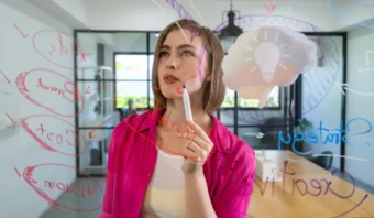 Una mujer con una camisa rosa escribe en una pizarra de cristal cubierta con diagramas y textos coloridos. Al fondo se ve el dibujo de una bombilla.