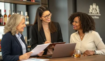 Tres mujeres vestidas de negocios colaboran en una mesa con documentos, una computadora portátil y una bebida en un entorno de oficina moderno.