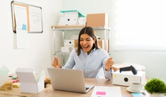 Una mujer está sentada en un escritorio en una pequeña oficina, sonriendo y levantando los puños con entusiasmo mientras mira una computadora portátil. A su alrededor se ven cajas de envío y materiales de embalaje.