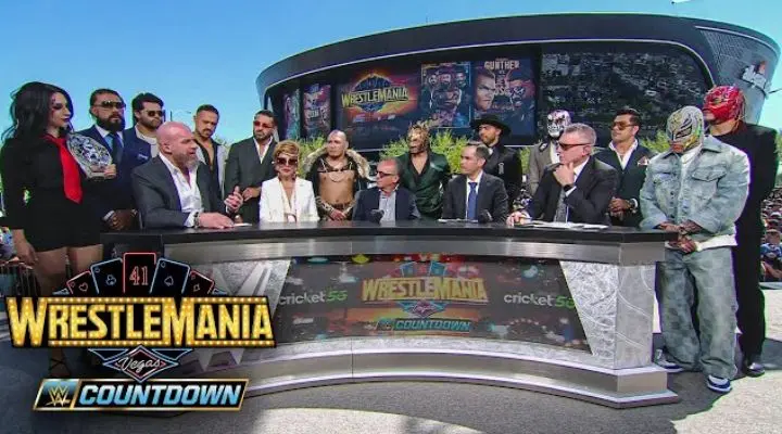Un grupo de personalidades de la WWE se sientan y se paran alrededor de un gran escritorio durante una transmisión de WrestleMania 41 Countdown afuera de una arena, con micrófonos y gráficos promocionales visibles.