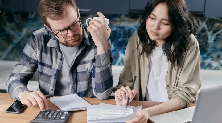 Un hombre y una mujer se sientan a una mesa revisando documentos con una calculadora y una computadora portátil, hablando de los efectos de la contracción inflacionaria. El hombre sostiene una taza mientras la mujer escribe con un bolígrafo, ambos concentrados en encontrar soluciones ante la shrinkflation y las cambiantes tendencias económicas.