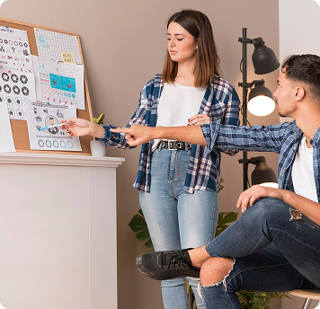 Dos personas discuten gráficos de datos en un tablero de corcho; una está de pie señalando un gráfico, la otra está sentada y hace un gesto hacia el tablero.