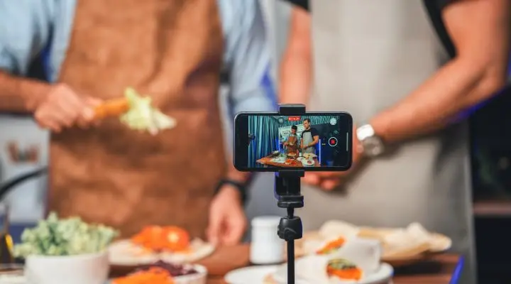 Dos personas se dedican al marketing gastronómico mientras cocinan y se graban con un smartphone montado en un trípode. Se ven diversos ingredientes en la mesa, creando un espectáculo visual encantador, perfecto para atraer a los amantes de la cocina.