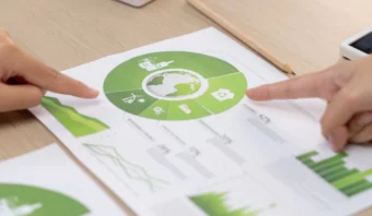 Dos manos señalan una infografía circular verde sobre un papel, que representa conceptos de sostenibilidad ambiental, con gráficos y cuadros en el fondo.