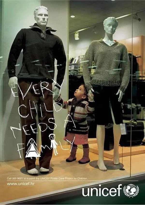Un niño se toma de la mano de dos maniquíes en el escaparate de una tienda, destacando los anuncios creativos. Un texto manuscrito dice: «Todo niño necesita una familia». El logotipo y el sitio web de UNICEF aparecen en la parte inferior.