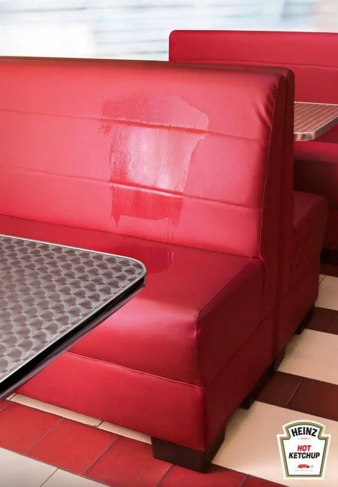 Una cabina de comedor roja con la forma de una botella de ketchup delineada en un tono más claro en el asiento, al lado de un logotipo de Heinz Hot Ketchup en la esquina, perfecta para anuncios creativos que llamen la atención.