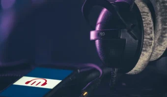 Un par de auriculares negros supraaurales etiquetados "PRO" descansan junto a un teléfono inteligente que muestra una aplicación de música con un ícono rojo y blanco, perfecto para sintonizar su podcast b2b favorito mientras viaja.