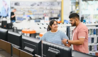Dos personas están juntas en una tienda de electrónica, explorando los televisores en los estantes. El fondo resalta la tecnología con diversos aparatos electrónicos y elementos de la tienda desenfocados.