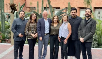 Siete personas están de pie en fila al aire libre frente a grandes plantas y un edificio de ladrillos, posando para una foto grupal como si estuvieran a punto de revelar El secreto del oro.