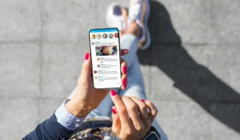 Una persona con uñas esmaltadas de color rosa sostiene un teléfono inteligente que muestra los medios en una red social mientras está parada sobre una superficie pavimentada.