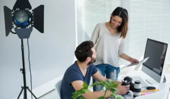 Un hombre sentado en un escritorio con una cámara habla con una mujer que sostiene papeles en una oficina con una computadora, una lente de cámara y una luz de estudio: un espacio de trabajo ideal para creadores de contenido.