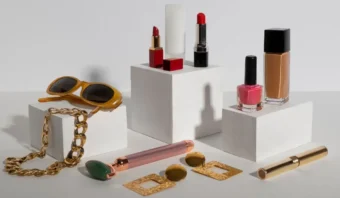 Cosméticos variados, gafas de sol, joyas de oro y un rodillo facial (bienes clásicos de Veblen) expuestos en bloques blancos sobre un fondo claro.