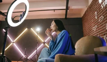Una mujer se sienta en un sofá comiendo pizza frente a un anillo de luz y una cámara, creando contenido para impulsar la reputación de marca con personas influyentes, con iluminación de estudio y una pared de ladrillos al fondo.