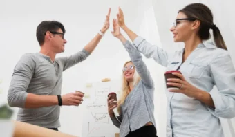 Tres personas vestidas de manera informal chocan los cinco y sonríen en un entorno de oficina, sosteniendo tazas de café y con una pizarra y un gráfico de fondo, celebrando el trabajo en equipo y la ética en el trabajo.