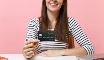 Una mujer que viste una camisa a rayas sostiene una tarjeta de crédito negra en su mano, sentada en un escritorio con un fondo rosa.